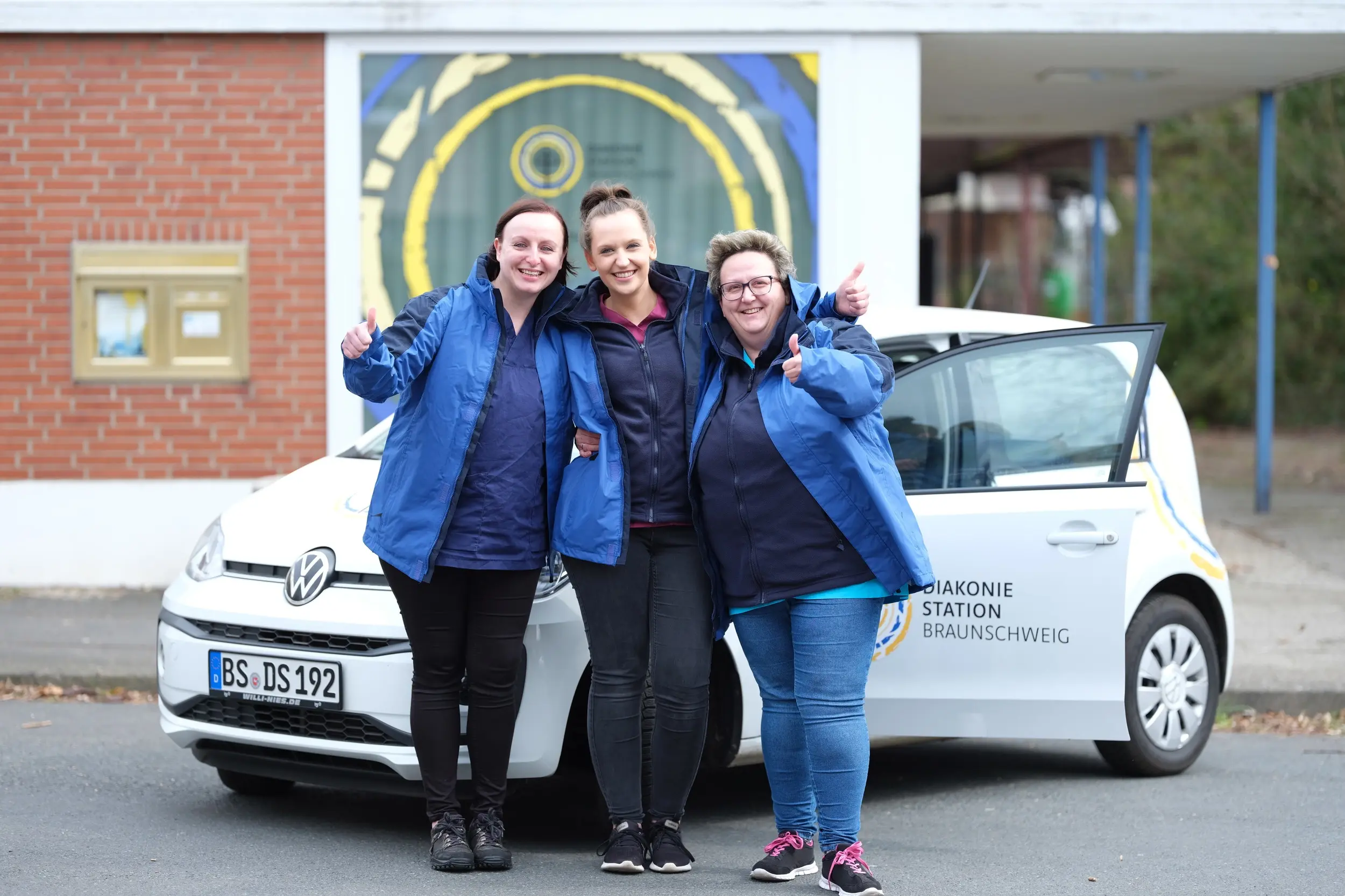 Fröhliches Pflegeteam der ambulanten Pflege steht am Auto und lacht in die Kamera
