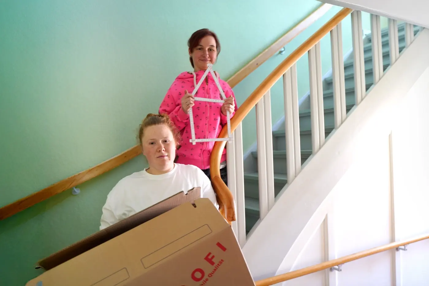 Zwei junge Menschen auf einer Treppe. eine zeigt ein Haus aus einem Zollstock gelegt, die andere hält einen Umzugskarton
