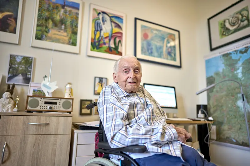 Alter Mann sitzt in seinem Zimmer im Rollstuhl, umgeben von Kunstwerken an den Wänden.