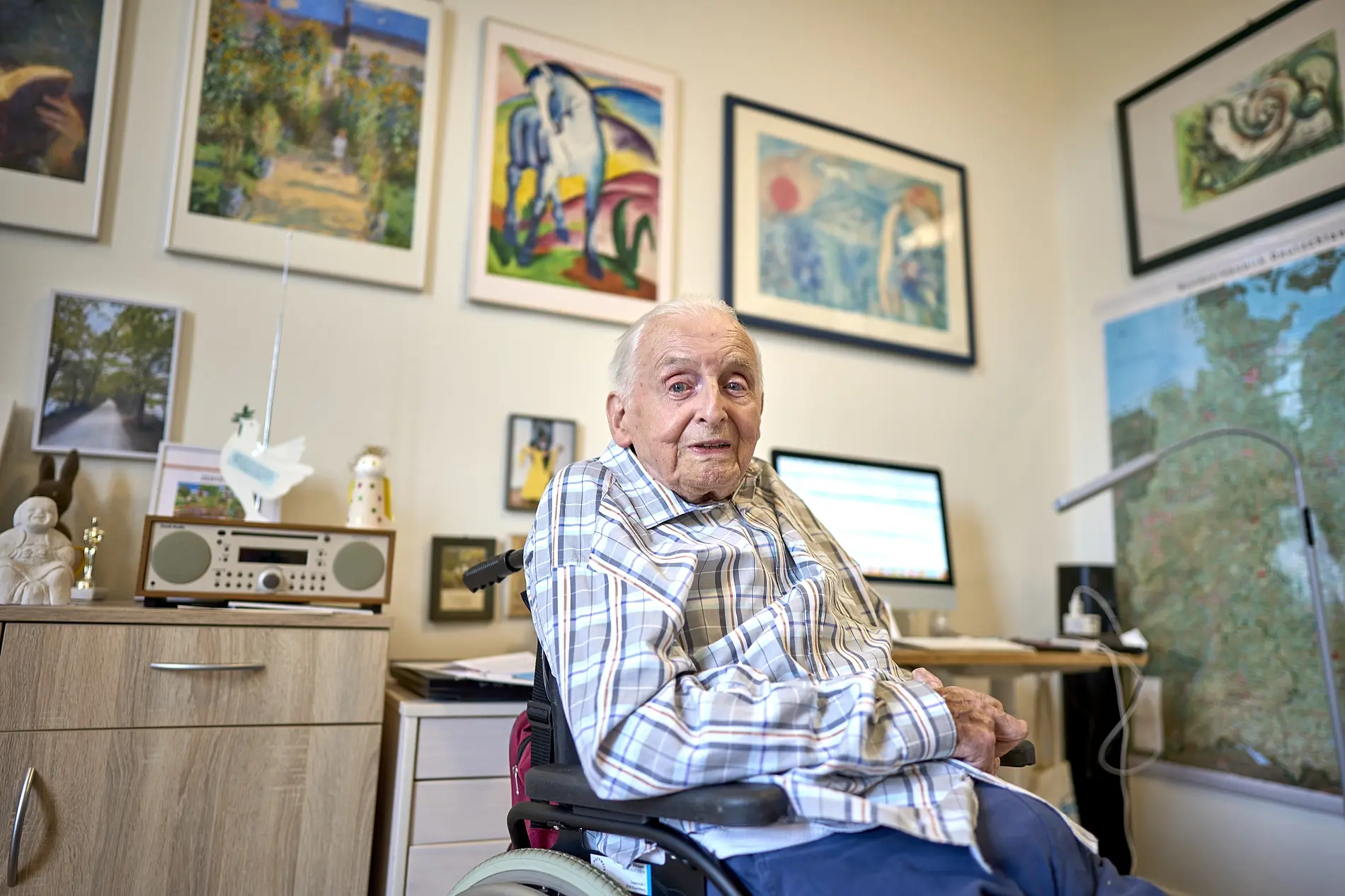 Alter Mann sitzt in seinem Zimmer im Rollstuhl, umgeben von Kunstwerken an den Wänden.