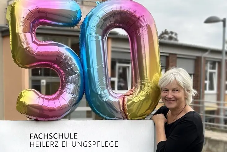 Annegret Jäkel steht neben dem Schild der Fachschule Heilerziehungspflege und hält einen Ballon mit einer großen, bunten 50