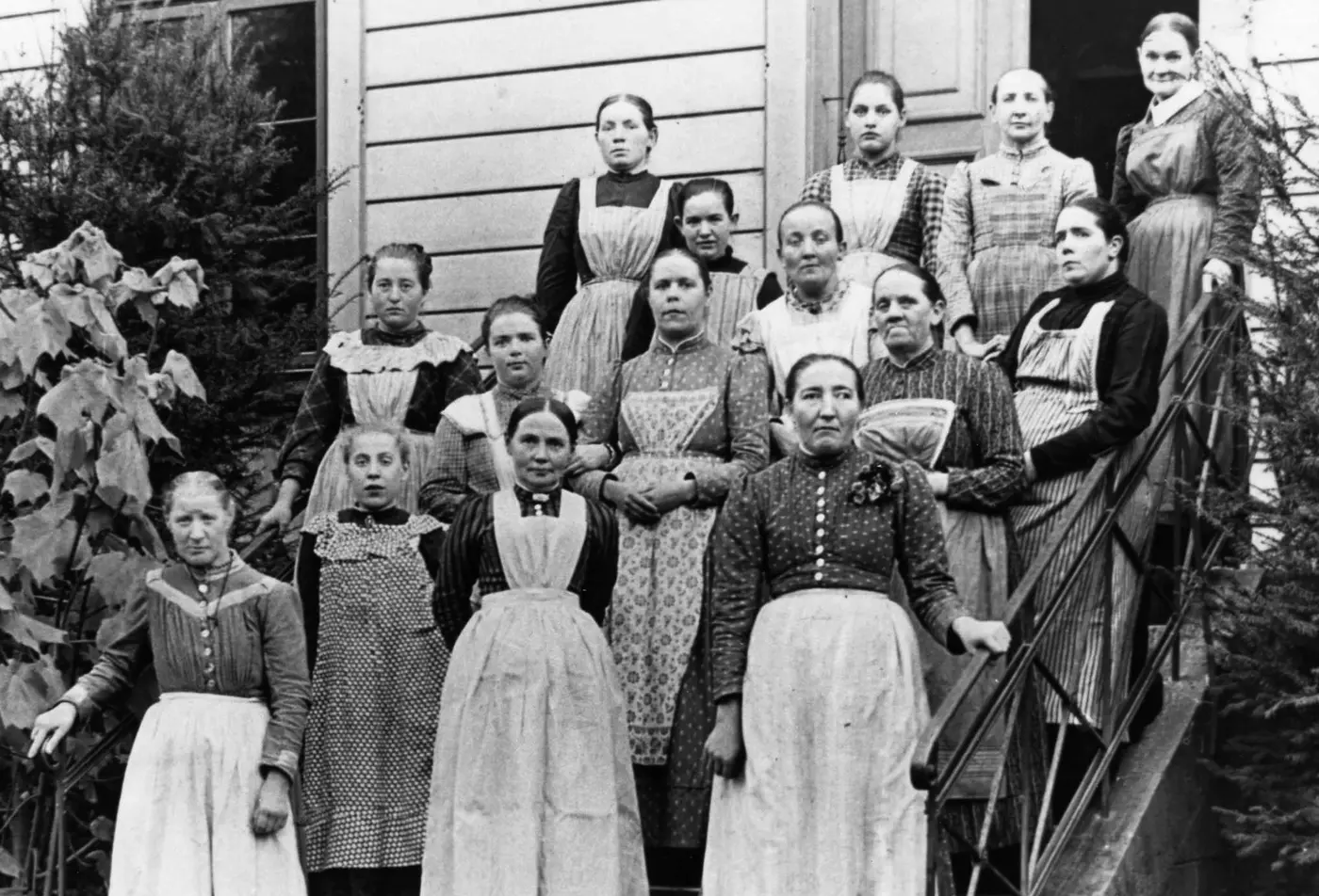 Historisches Bild von 1912. Frauen mit Behinderungen stehen in Kleidern und weißen Schürzen vor auf einer Treppe.