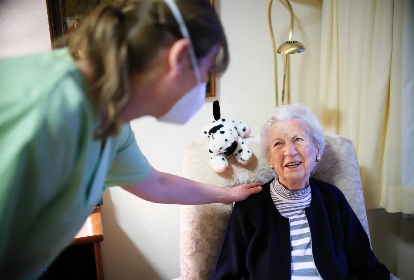 Pflegefachkraft betreut Seniorin in ihrem Zimmer, diese sitzt in einem Sessel beide lächeln sich an