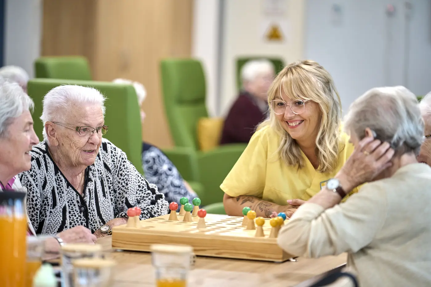 Das Bild zeigt eine Mitarbeiterin und Seniorinnen, die in einer Tagespflege Mensch-ärgere-dich-nicht spielen.