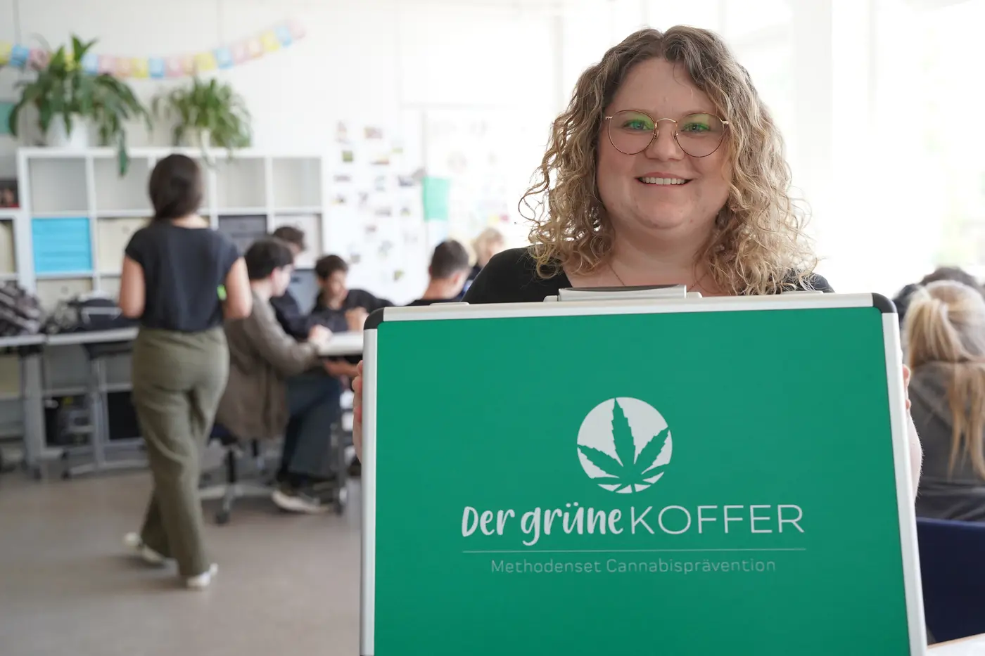 Frau mit lockigen blonden Haaren zeigt den "Grünen Koffer" in die Kamera