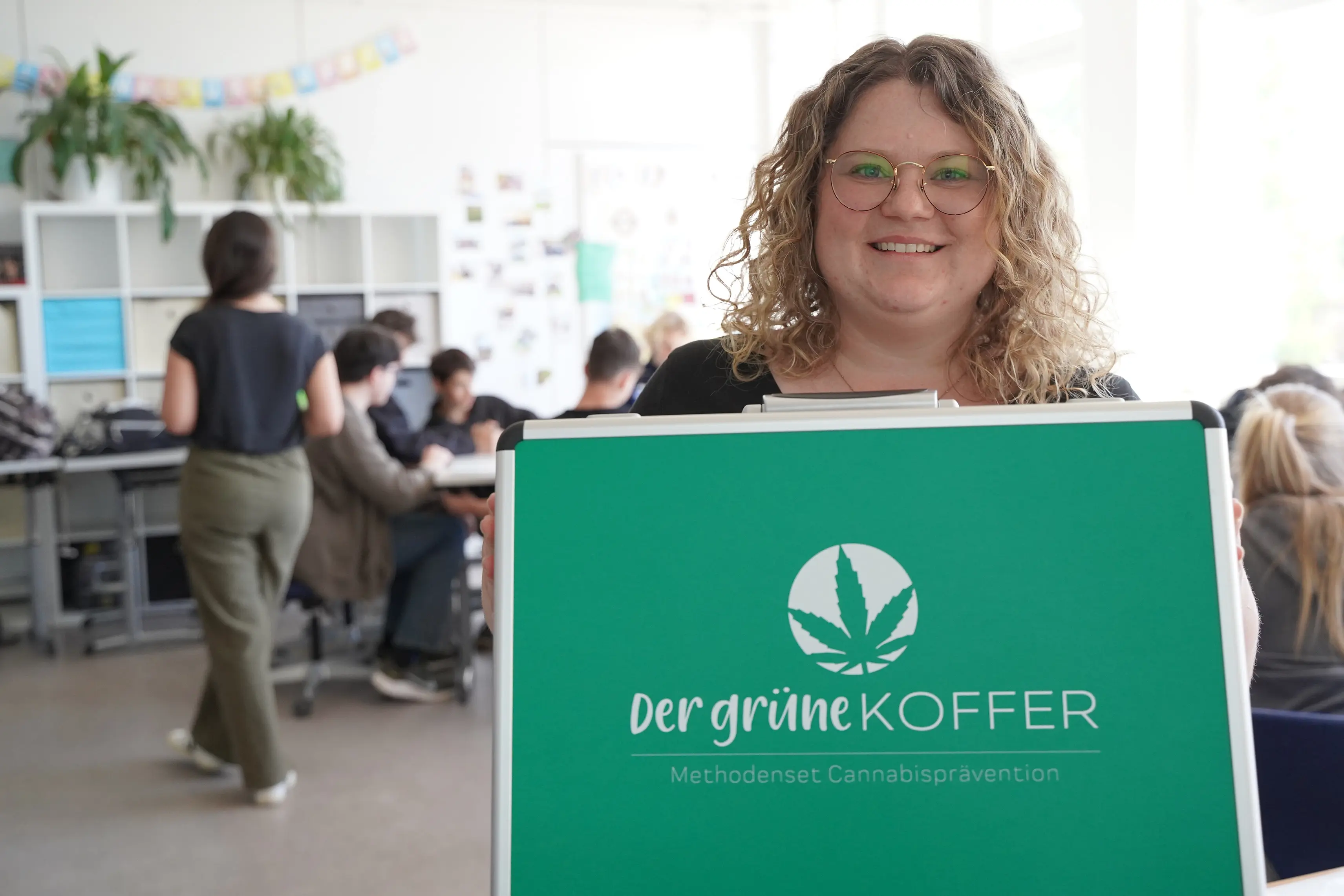 Frau mit lockigen blonden Haaren zeigt den "Grünen Koffer" in die Kamera